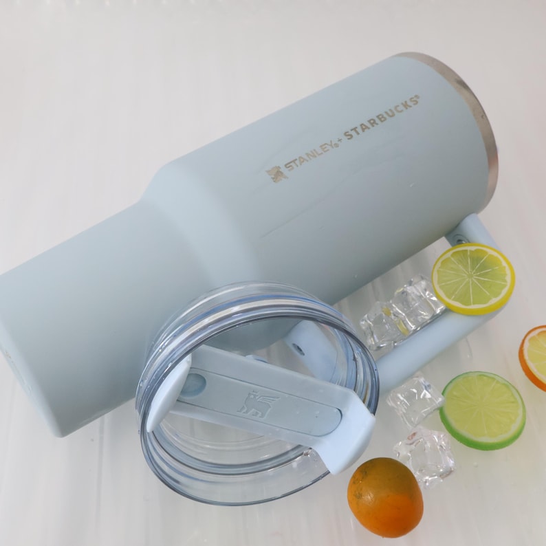 Puede incluir: Un vaso Stanley Starbucks azul claro con tapa transparente y asa, con el nombre de la marca impreso en el lateral. El vaso est&aacute; junto a rodajas de lim&oacute;n y lima, cubitos de hielo y una peque&ntilde;a naranja.