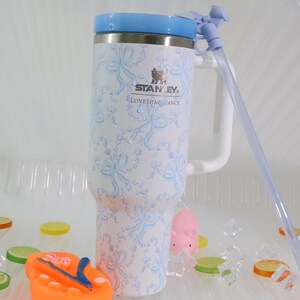 K&ouml;nnte beinhalten: Ein wei&szlig;-blauer Stanley Loveshack Fancy Becher mit Griff und Strohhalm. Der Becher hat einen hellblauen Deckel und ein Muster aus blauen Schleifen. Der Becher hat das Stanley-Logo und die Aufschrift "Loveshack Fancy".