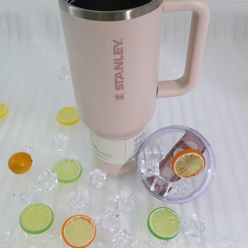 Peut inclure: Un mug Stanley rose clair avec une poign&eacute;e et un bord argent&eacute;, portant le nom de la marque. Le mug est &agrave; c&ocirc;t&eacute; d'un couvercle transparent avec une tranche d'orange &agrave; l'int&eacute;rieur, entour&eacute; de gla&ccedil;ons et de tranches d'agrumes d&eacute;coratives.