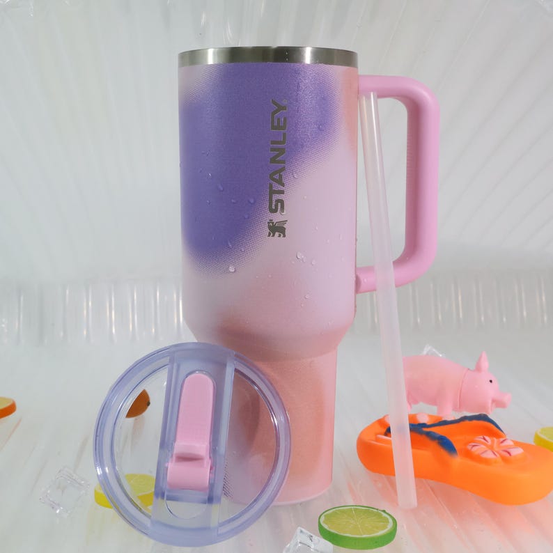 Puede incluir: Un vaso aislado Stanley en degradado de morado y rosa, con asa y tapa rosas a juego. El vaso tiene el logotipo de Stanley impreso en el lateral. Se incluye una pajita y una tapa transparentes.