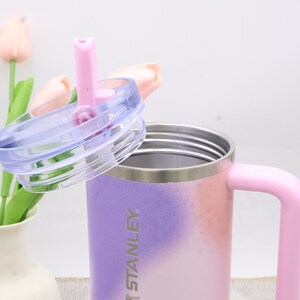 Puede incluir: Un vaso t&eacute;rmico Stanley con asa rosa y un dise&ntilde;o degradado de rosa, morado y blanco. La tapa transparente tiene una pajita rosa. El vaso est&aacute; dise&ntilde;ado para mantener las bebidas calientes o fr&iacute;as.