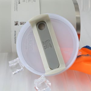 May include: Close-up of a white and gray Stanley Quencher lid. The lid features a gray slider with the Stanley logo and a small circular opening. The lid is partially open, revealing the interior. Ice cubes and an orange and blue object are in the background.