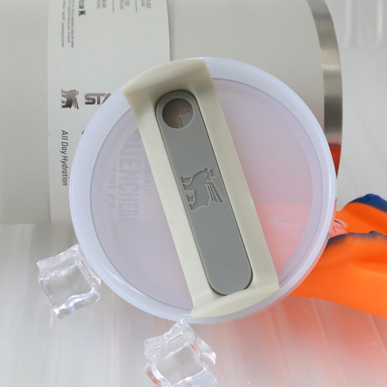 May include: Close-up of a white and gray Stanley Quencher lid. The lid features a gray slider with the Stanley logo and a small circular opening. The lid is partially open, revealing the interior. Ice cubes and an orange and blue object are in the background.