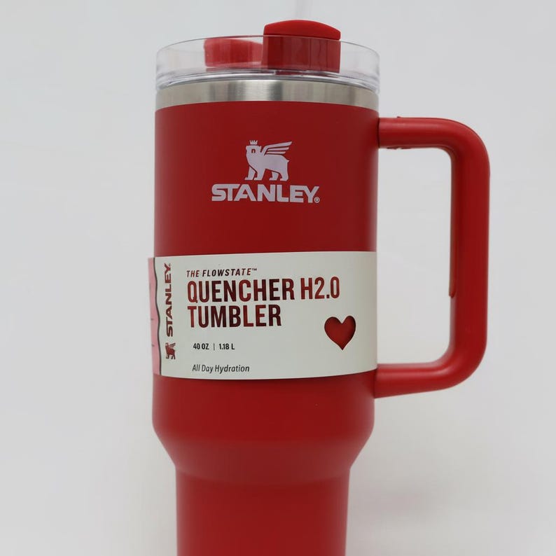 Może przedstawiać: Czerwony kubek Stanley Quencher H2.0 z uchwytem i przezroczystą pokrywką. Kubek ma logo Stanley i napis "QUENCHER H2.0 TUMBLER" i mieści 1,18 L.
