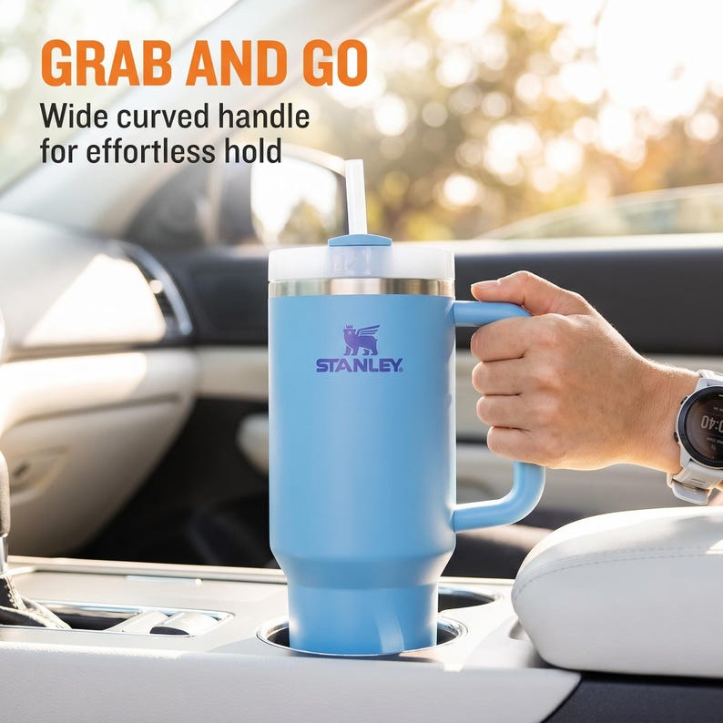 May include: A light blue Stanley travel mug with a white lid and straw, held in a car's cup holder. The mug has a curved handle and the Stanley logo. Text on the image reads "GRAB AND GO" and "Wide curved handle for effortless hold."
