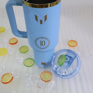 May include: A light blue insulated tumbler with a gold rim and handle. The tumbler has a logo and the number 10 printed on it. The lid is clear with a lime slice inside. Decorative ice cubes and fruit slices surround the tumbler.