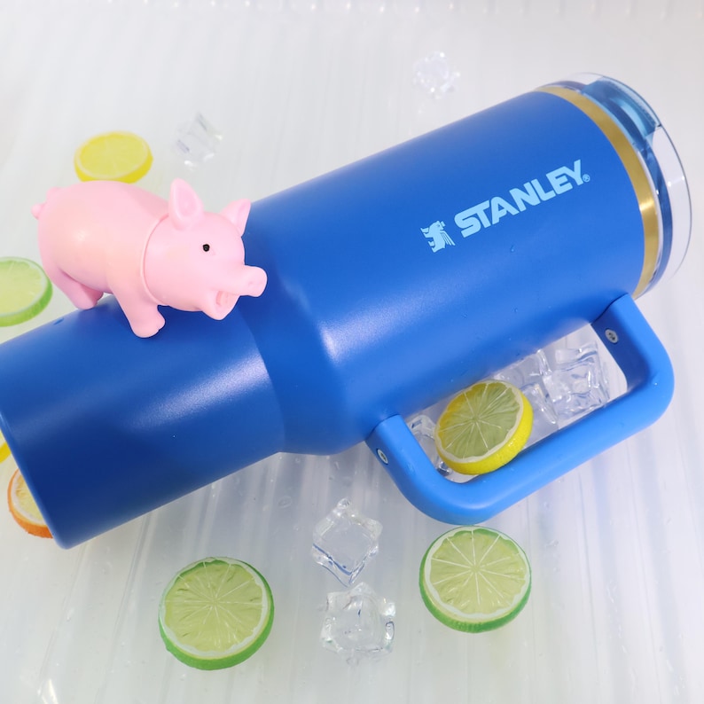 May include: A blue Stanley tumbler with a handle, featuring the brand name in white. A small pink pig figurine rests on the tumbler. The scene includes lemon and lime slices, and ice cubes, suggesting a refreshing drink.