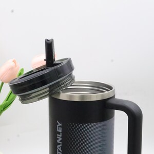Puede incluir: Taza de viaje aislada negra y plateada con asa y tapa negra. La taza tiene la palabra "STANLEY" impresa en el lateral. La tapa est&aacute; parcialmente abierta, revelando el interior de la taza.