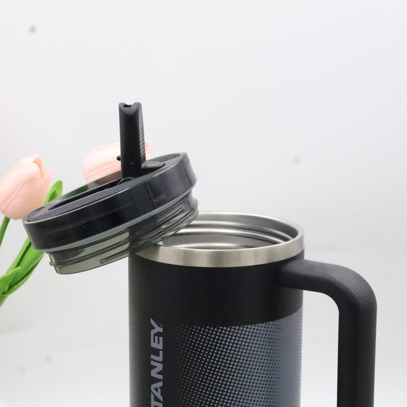 Puede incluir: Taza de viaje aislada negra y plateada con asa y tapa negra. La taza tiene la palabra "STANLEY" impresa en el lateral. La tapa est&aacute; parcialmente abierta, revelando el interior de la taza.