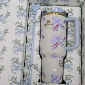May include: A white Stanley Quencher tumbler with a floral and bow pattern in purple and pink, with a light blue handle and lid. The tumbler is in a matching patterned box with a light blue straw.