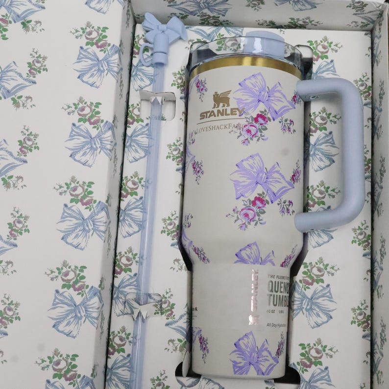 May include: A white Stanley Quencher tumbler with a floral and bow pattern in purple and pink, with a light blue handle and lid. The tumbler is in a matching patterned box with a light blue straw.