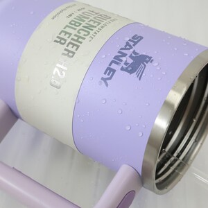 Może przedstawiać: Liliowy kubek Stanley Quencher H2.0 ze stalową obręczą i uchwytem. Kubek ma logo Stanley i informacje o produkcie. Widoczne są krople wody.