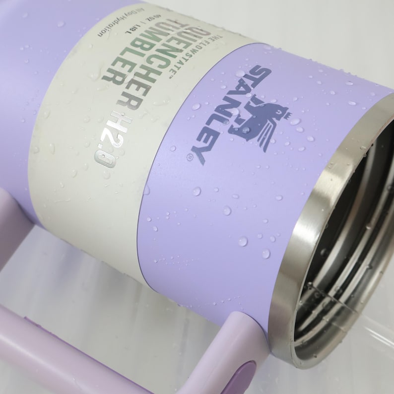 Może przedstawiać: Liliowy kubek Stanley Quencher H2.0 ze stalową obręczą i uchwytem. Kubek ma logo Stanley i informacje o produkcie. Widoczne są krople wody.
