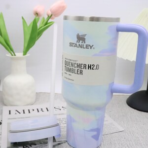 May include: A Stanley Quencher H2.0 tumbler with a light blue and purple marbled design. The tumbler has a light purple handle and the text "QUENCHER H2.0 TUMBLER". The tumbler is designed for 40-day hydration.