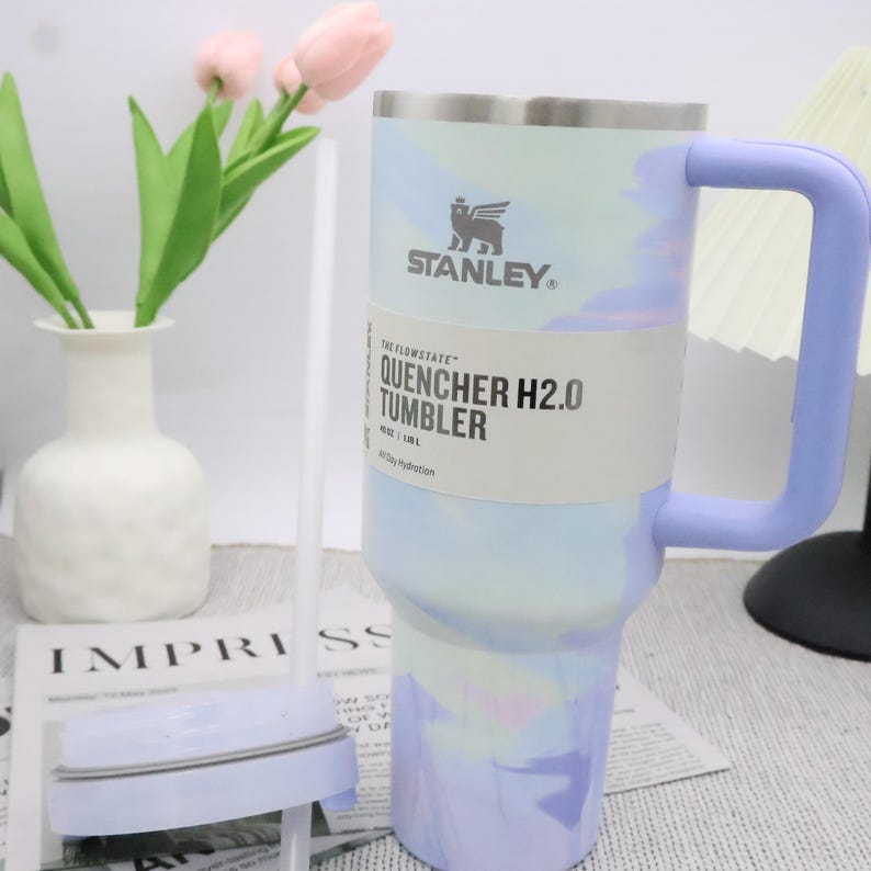 May include: A Stanley Quencher H2.0 tumbler with a light blue and purple marbled design. The tumbler has a light purple handle and the text "QUENCHER H2.0 TUMBLER". The tumbler is designed for 40-day hydration.