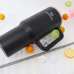 May include: A black Stanley tumbler with a handle, ice cubes, and a straw. The tumbler has the Stanley logo and is surrounded by oranges and lime slices. The tumbler is designed to keep drinks cold.