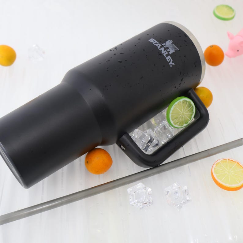 May include: A black Stanley tumbler with a handle, ice cubes, and a straw. The tumbler has the Stanley logo and is surrounded by oranges and lime slices. The tumbler is designed to keep drinks cold.