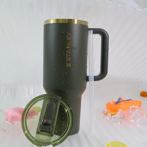 Puede incluir: Un termo de viaje Stanley verde con borde dorado y asa negra. El termo tiene el logotipo de Stanley impreso en el lateral. La tapa es verde transparente. El termo est&aacute; dise&ntilde;ado para mantener las bebidas calientes o fr&iacute;as.