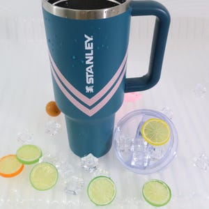 K&ouml;nnte beinhalten: Ein tealfarbener Stanley-Reisebecher mit Griff und silbernem Rand. Der Becher hat ein rosa-wei&szlig;es Chevron-Design und das Stanley-Logo. Ein klarer Deckel mit einer Zitronenscheibe befindet sich neben dem Becher, zusammen mit Eisw&uuml;rfeln und Zitronenscheiben.
