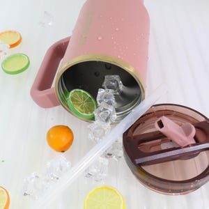 May include: A pink insulated tumbler with a handle, gold trim, and a brown lid. The tumbler is partially filled with ice cubes and a lime slice. A clear straw is next to the tumbler. Decorative fruit slices are scattered around the tumbler.