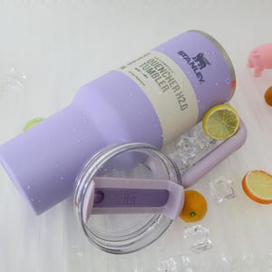 Puede incluir: Un vaso Stanley Quencher H2.0 de color lila claro con tapa transparente y asa. El vaso tiene el texto "QUENCHER H2.0 TUMBLER" impreso. La imagen tambi&eacute;n incluye cubitos de hielo, rodajas de lim&oacute;n y naranjas peque&ntilde;as.