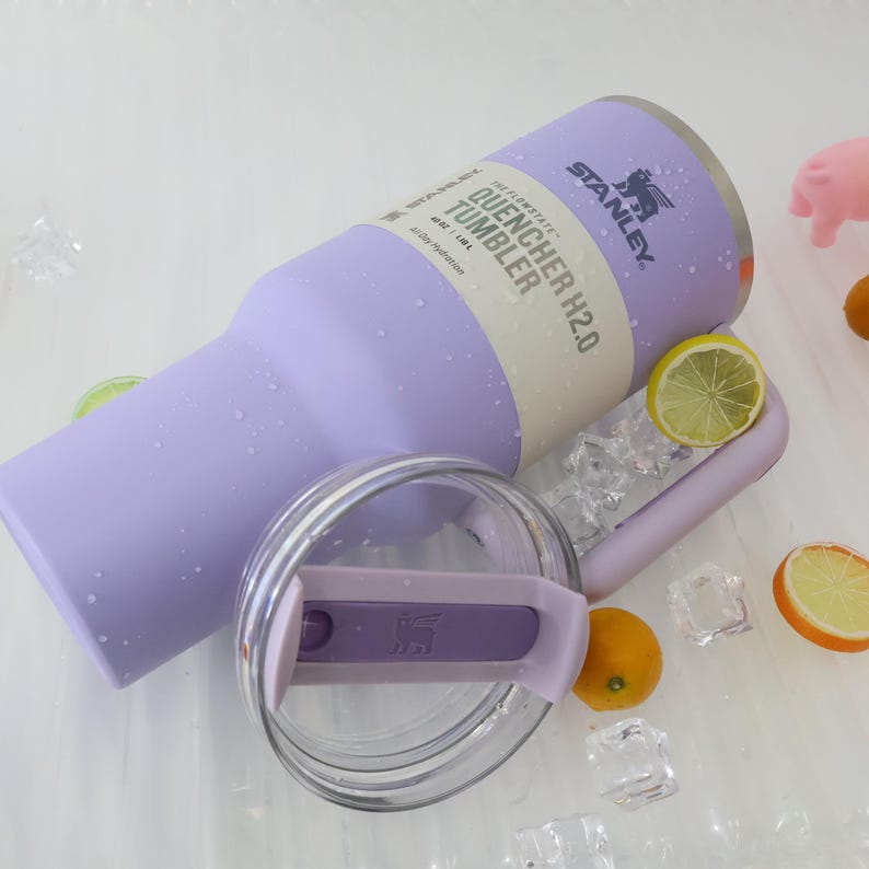 Puede incluir: Un vaso Stanley Quencher H2.0 de color lila claro con tapa transparente y asa. El vaso tiene el texto "QUENCHER H2.0 TUMBLER" impreso. La imagen tambi&eacute;n incluye cubitos de hielo, rodajas de lim&oacute;n y naranjas peque&ntilde;as.
