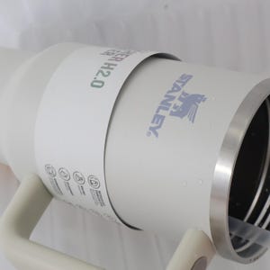 May include: A white Stanley H2.0 water bottle with a silver rim and handle. The Stanley logo is printed in gray. The bottle has a white paper band wrapped around it with the text "WATER H2.0" in green.