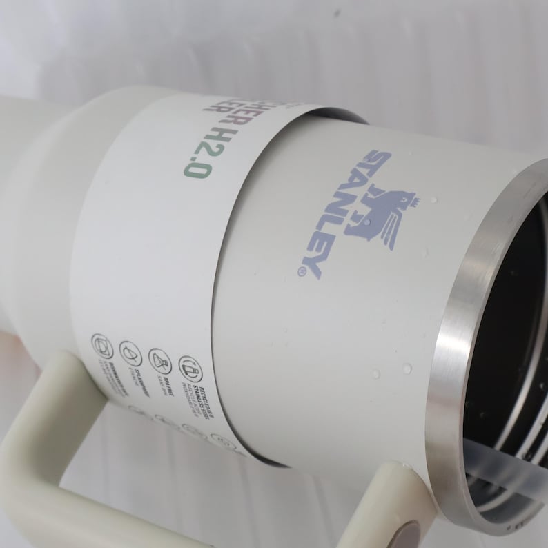 May include: A white Stanley H2.0 water bottle with a silver rim and handle. The Stanley logo is printed in gray. The bottle has a white paper band wrapped around it with the text "WATER H2.0" in green.
