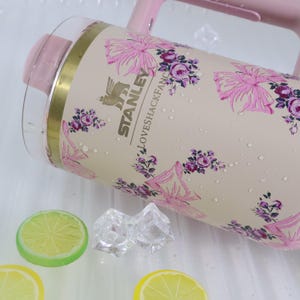 K&ouml;nnte beinhalten: Ein rosa-transparenter Stanley-Wasserkrug mit floralem Design. Der Krug hat einen hellbeigen Hintergrund mit rosa Blumen und dem Text "LOVESHACKFANCY". Der Krug hat einen rosa Griff und Deckel. Er liegt auf einer wei&szlig;en Oberfl&auml;che mit Zitronenscheiben und Eisw&uuml;rfeln.