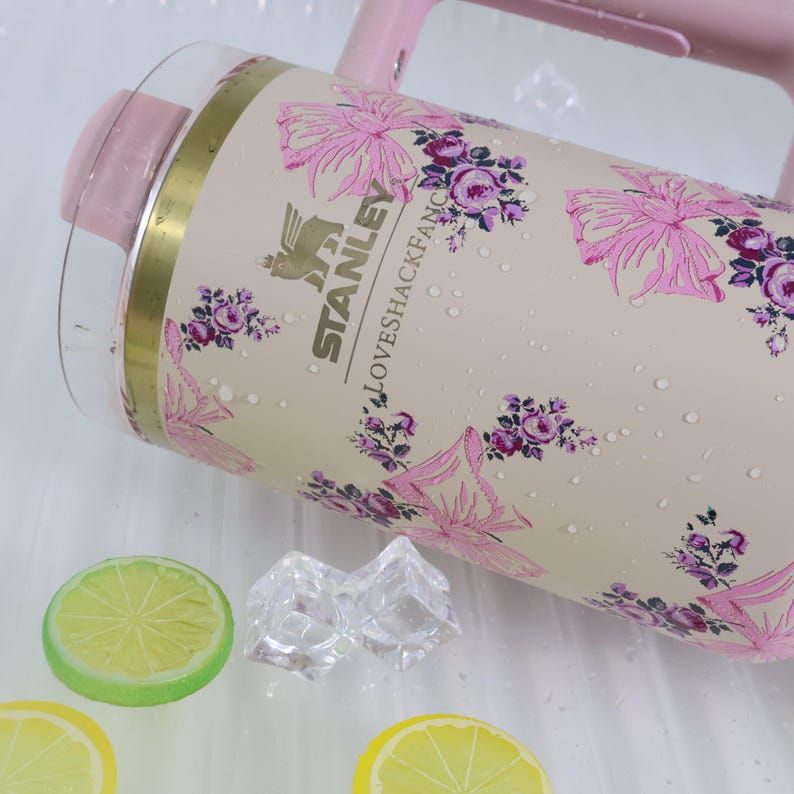 K&ouml;nnte beinhalten: Ein rosa-transparenter Stanley-Wasserkrug mit floralem Design. Der Krug hat einen hellbeigen Hintergrund mit rosa Blumen und dem Text "LOVESHACKFANCY". Der Krug hat einen rosa Griff und Deckel. Er liegt auf einer wei&szlig;en Oberfl&auml;che mit Zitronenscheiben und Eisw&uuml;rfeln.