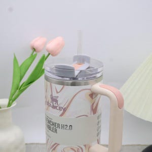 May include: A stainless steel tumbler with a marble pattern in shades of pink and brown. It has a clear lid, a pink handle, and a straw. The tumbler has the text "ENCHER H2.0 TUMBLER".