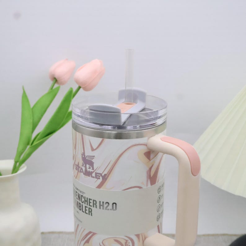 May include: A stainless steel tumbler with a marble pattern in shades of pink and brown. It has a clear lid, a pink handle, and a straw. The tumbler has the text "ENCHER H2.0 TUMBLER".
