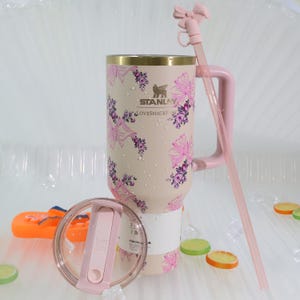 K&ouml;nnte beinhalten: Ein beigefarbener Stanley Loveshackfancy-Becher mit goldenem Rand, verziert mit rosa Schleifen und floralen Mustern. Er hat einen rosa Griff, Strohhalm und Deckel. Der Becher ist etwa 25 cm hoch.