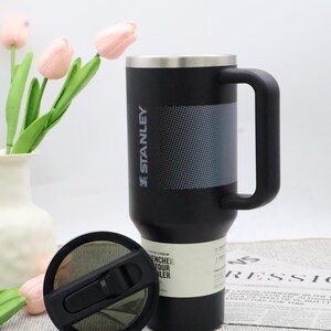 Puede incluir: Taza de viaje Stanley negra con borde plateado y asa negra grande. La taza tiene un panel gris texturizado y el logotipo de Stanley. La tapa es negra con una pajita abatible. Un jarr&oacute;n de tulipanes rosas est&aacute; en el fondo.