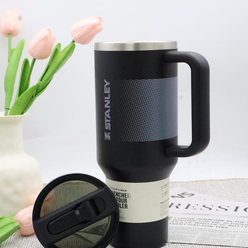 Puede incluir: Taza de viaje Stanley negra con borde plateado y asa negra grande. La taza tiene un panel gris texturizado y el logotipo de Stanley. La tapa es negra con una pajita abatible. Un jarr&oacute;n de tulipanes rosas est&aacute; en el fondo.