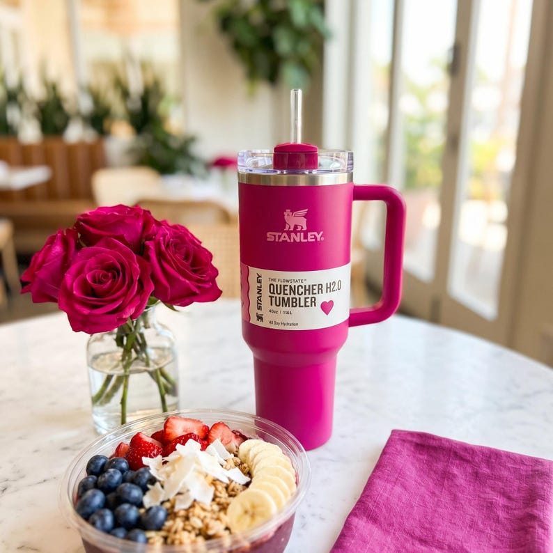 Peut inclure: Un gobelet Stanley Quencher H2.0 rose vif avec une poign&eacute;e et une paille, &agrave; c&ocirc;t&eacute; d'un bol de fruits et d'un bouquet de roses roses. Le gobelet porte l'inscription "QUENCHER H2.0 TUMBLER". Il contient 1,18L.