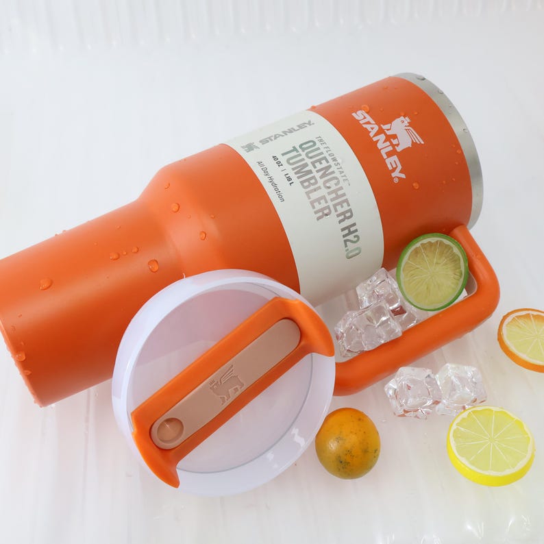 Puede incluir: Un vaso Stanley Quencher H2.0 naranja con tapa blanca y naranja. El vaso tiene un asa y el texto "QUENCHER H2.0 TUMBLER" es visible. La tapa est&aacute; quitada y hay cubitos de hielo y rodajas de lim&oacute;n cerca. Capacidad: 1,18 L.