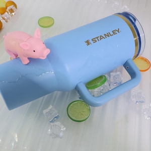 May include: A light blue Stanley tumbler with a handle, gold accents, and the brand name printed on the side. A small pink pig figurine sits on the tumbler. Decorative lime and orange slices and ice cubes are scattered around the tumbler.