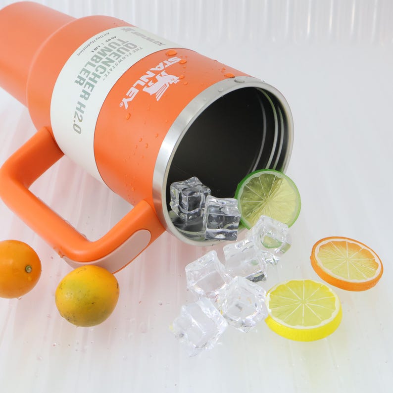 Puede incluir: Un vaso naranja Stanley Quencher H2.0 con asa, cubitos de hielo y rodajas de c&iacute;tricos. El vaso tiene el texto "QUENCHER TUMBLER H2.0" y el logotipo de Stanley. El vaso est&aacute; parcialmente lleno de cubitos de hielo y rodeado de rodajas de lim&oacute;n y lima.