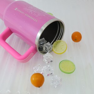 Puede incluir: Un vaso rosa Stanley Starbucks con asa, cubitos de hielo y rodajas de c&iacute;tricos. El vaso tiene el texto "STANLEY STARBUCKS" impreso en el lateral. Tambi&eacute;n hay dos naranjas peque&ntilde;as.