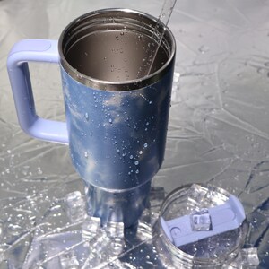 K&ouml;nnte beinhalten: Ein blau-silberner isolierter Reisebecher mit Griff und transparentem Deckel. Der Becher hat ein hellblaues Marmormuster und ist mit Wassertropfen bedeckt. Der Deckel ist transparent mit einem hellblauen Schieber.