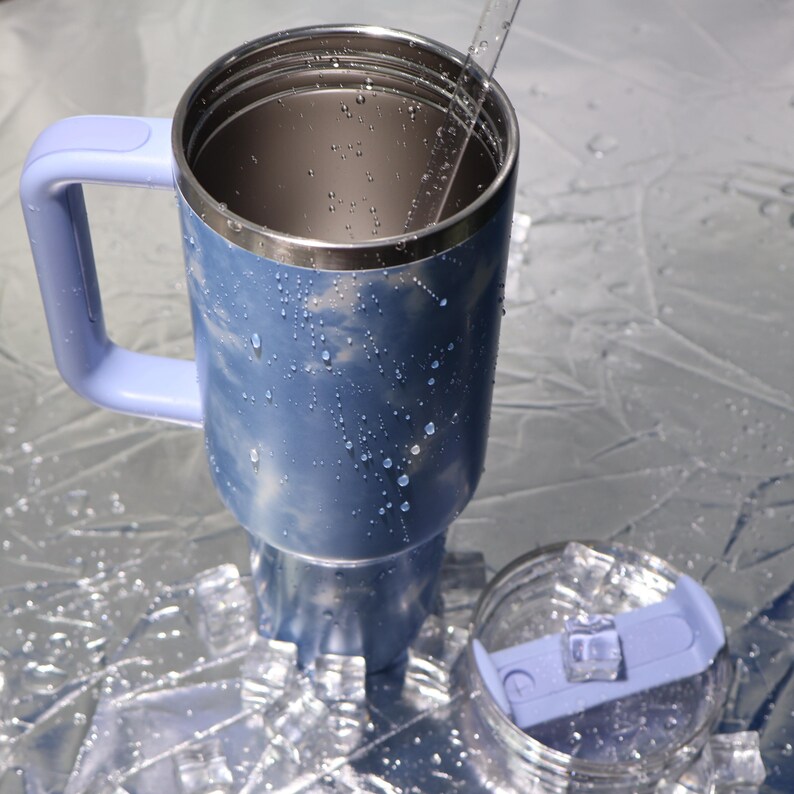 K&ouml;nnte beinhalten: Ein blau-silberner isolierter Reisebecher mit Griff und transparentem Deckel. Der Becher hat ein hellblaues Marmormuster und ist mit Wassertropfen bedeckt. Der Deckel ist transparent mit einem hellblauen Schieber.