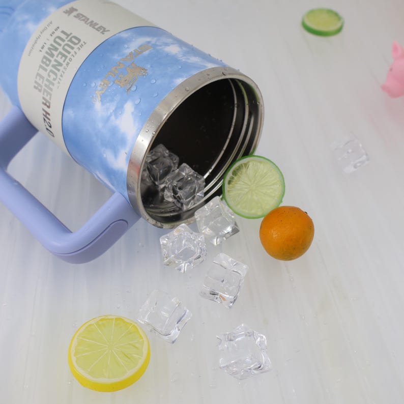 K&ouml;nnte beinhalten: Ein blau-wei&szlig;er Stanley Quencher H2.0 Becher mit lavendelfarbenem Griff, der auf der Seite liegt. Der Becher ist von Eisw&uuml;rfeln, einer Zitronenscheibe, einer Limettenscheibe und einer kleinen Orange umgeben. Der Becher tr&auml;gt den Text "QUENCHER TUMBLER H2.0".
