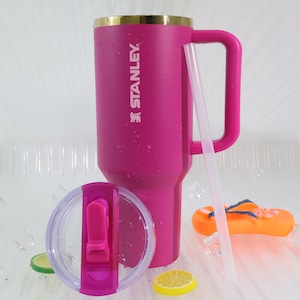 May include: A vibrant pink Stanley tumbler with a gold rim, a large handle, and a clear lid. A clear straw rests against the tumbler. The Stanley logo is printed on the side. The tumbler is next to a pair of orange flip-flops and lemon slices.