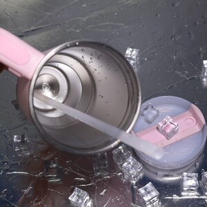K&ouml;nnte beinhalten: Ein Edelstahlbecher mit rosa Griff und Deckel sowie einem durchsichtigen Strohhalm. Der Deckel ist hellblau und rosa. Eisw&uuml;rfel umgeben den Becher auf einer reflektierenden Oberfl&auml;che.