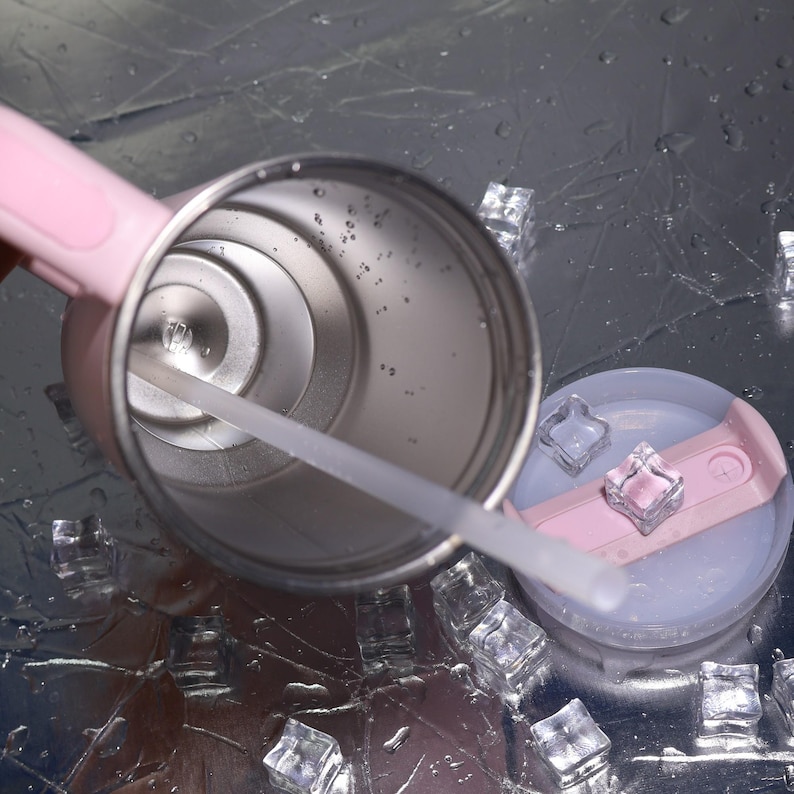 K&ouml;nnte beinhalten: Ein Edelstahlbecher mit rosa Griff und Deckel sowie einem durchsichtigen Strohhalm. Der Deckel ist hellblau und rosa. Eisw&uuml;rfel umgeben den Becher auf einer reflektierenden Oberfl&auml;che.