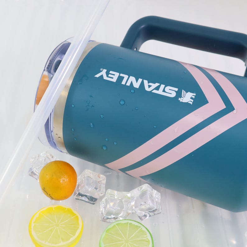 K&ouml;nnte beinhalten: Teal Stanley Wasserkanne mit rosa Chevron-Design und dunkelblauem Griff. Auf der Seite ist das Stanley-Logo aufgedruckt. Das Bild enth&auml;lt auch Eisw&uuml;rfel, eine Zitrone, eine Limette und eine Orange.