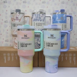 May include: Several Stanley Quencher H2.0 tumblers in pastel shades of pink, blue, and lilac. Each tumbler has a handle, lid, and the Stanley logo. The label displays the text "QUENCHER H2.0 TUMBLER". Measurements are in centimeters.