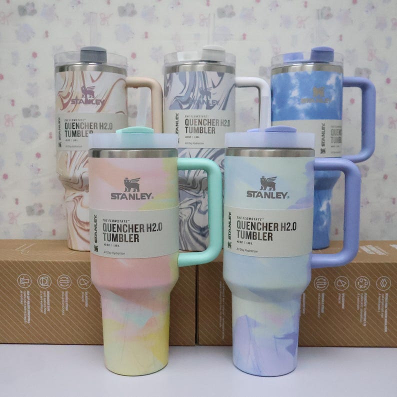 May include: Several Stanley Quencher H2.0 tumblers in pastel shades of pink, blue, and lilac. Each tumbler has a handle, lid, and the Stanley logo. The label displays the text "QUENCHER H2.0 TUMBLER". Measurements are in centimeters.