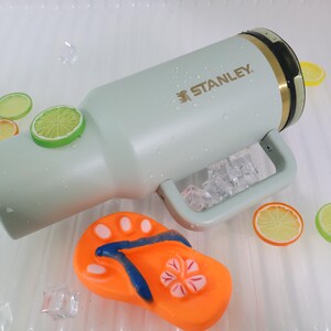 Op de afbeelding: Een lichtgroene Stanley waterfles met een handvat, ijsblokjes en het Stanley-logo. De fles is versierd met citroen- en limoenschijfjes en een oranje teenslipper.